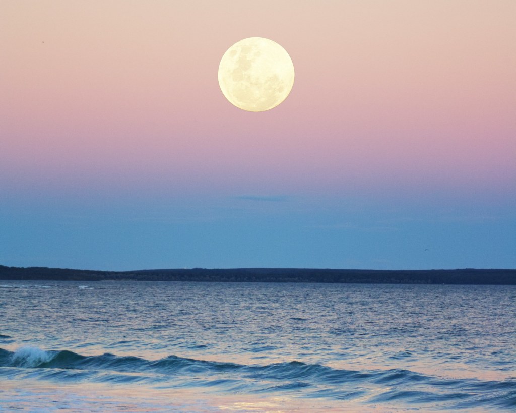 Full Moon even with light, sky degraded in shades of blue, purple, gold, pink, yellow. Calm sea.