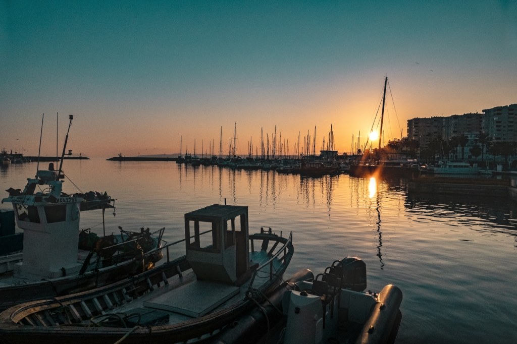 El sol poniéndose detrás de los mástiles de los barcos atracados, pintando el puerto con tonos dorados y naranjas.